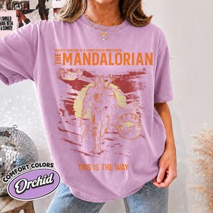 May include: An orchid-colored t-shirt featuring a graphic print of The Mandalorian. The design includes the text "Bounty hunting is a complicated profession," "The Mandalorian," and "This is the way" in orange, with a yellow sun background.