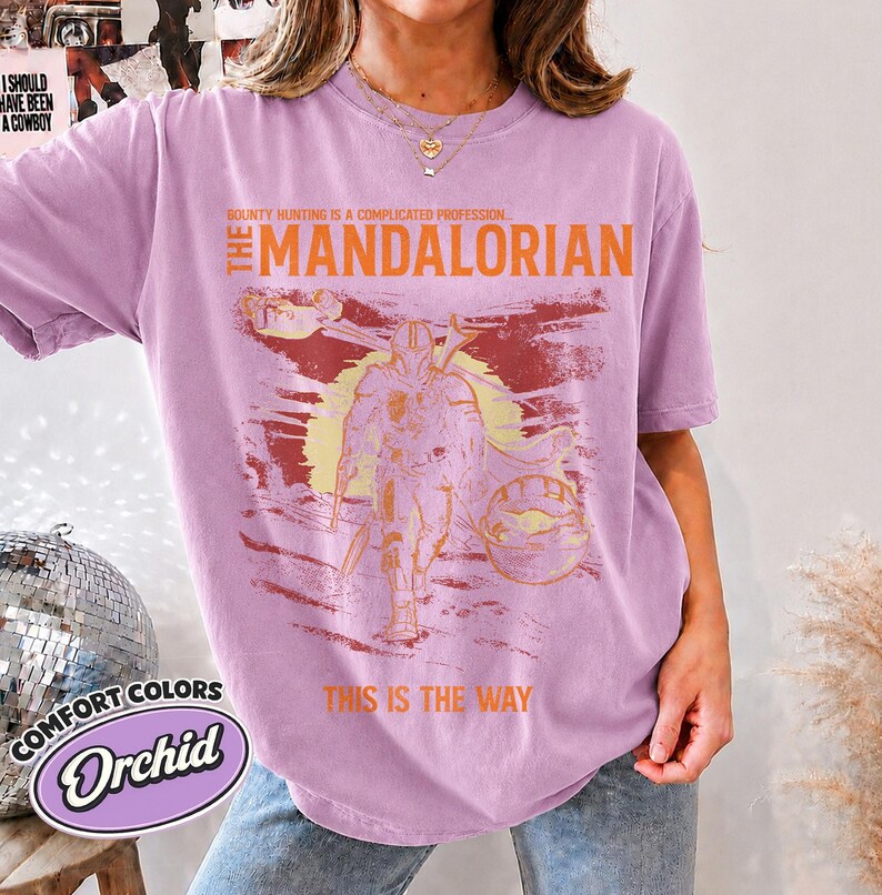 May include: An orchid-colored t-shirt featuring a graphic print of The Mandalorian. The design includes the text "Bounty hunting is a complicated profession," "The Mandalorian," and "This is the way" in orange, with a yellow sun background.