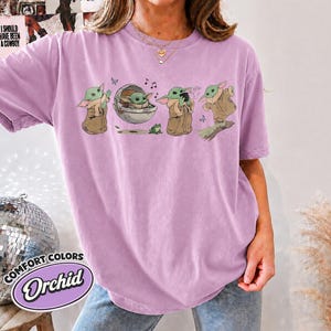 May include: A light purple short-sleeve t-shirt featuring a graphic print of a green creature in various poses, including sitting in a floating pod, holding a cup, and standing. The shirt has a crew neckline and a relaxed, loose-fitting silhouette.