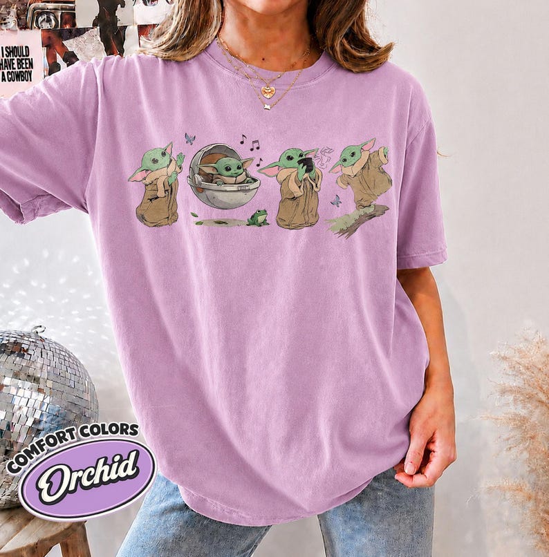 May include: A light purple short-sleeve t-shirt featuring a graphic print of a green creature in various poses, including sitting in a floating pod, holding a cup, and standing. The shirt has a crew neckline and a relaxed, loose-fitting silhouette.