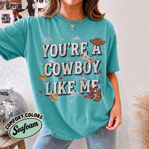 May include: A teal short-sleeve t-shirt featuring the text "YOU'RE A COWBOY LIKE ME" in white with a dark outline. The graphic includes a brown cowboy hat, a rocket ship, a gold sheriff star, a ball, and brown cowboy boots with red stars.