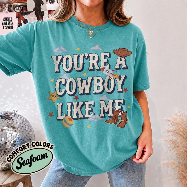 May include: A teal short-sleeve t-shirt featuring the text "YOU'RE A COWBOY LIKE ME" in white with a dark outline. The graphic includes a brown cowboy hat, a rocket ship, a gold sheriff star, a ball, and brown cowboy boots with red stars.