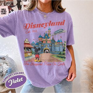 May include: A violet short-sleeve t-shirt featuring a graphic of a castle, a monorail, and a cartoon mouse. The text reads Disneyland Est. 1955 and The Happiest Place On Earth. The shirt has a crew neckline and a relaxed, vintage-style fit.