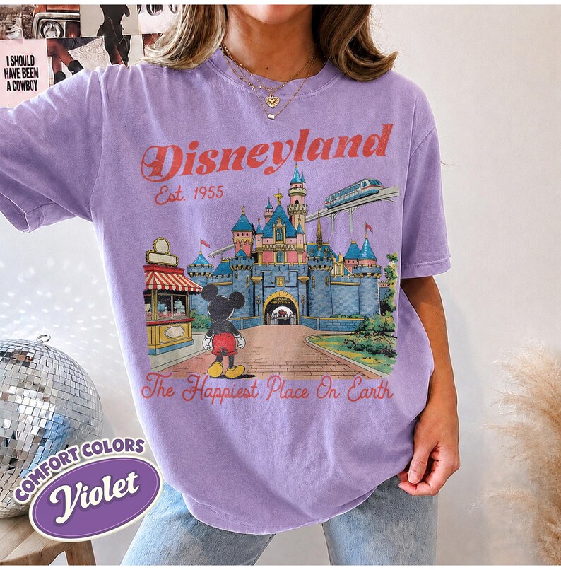 May include: A violet short-sleeve t-shirt featuring a graphic of a castle, a monorail, and a cartoon mouse. The text reads Disneyland Est. 1955 and The Happiest Place On Earth. The shirt has a crew neckline and a relaxed, vintage-style fit.