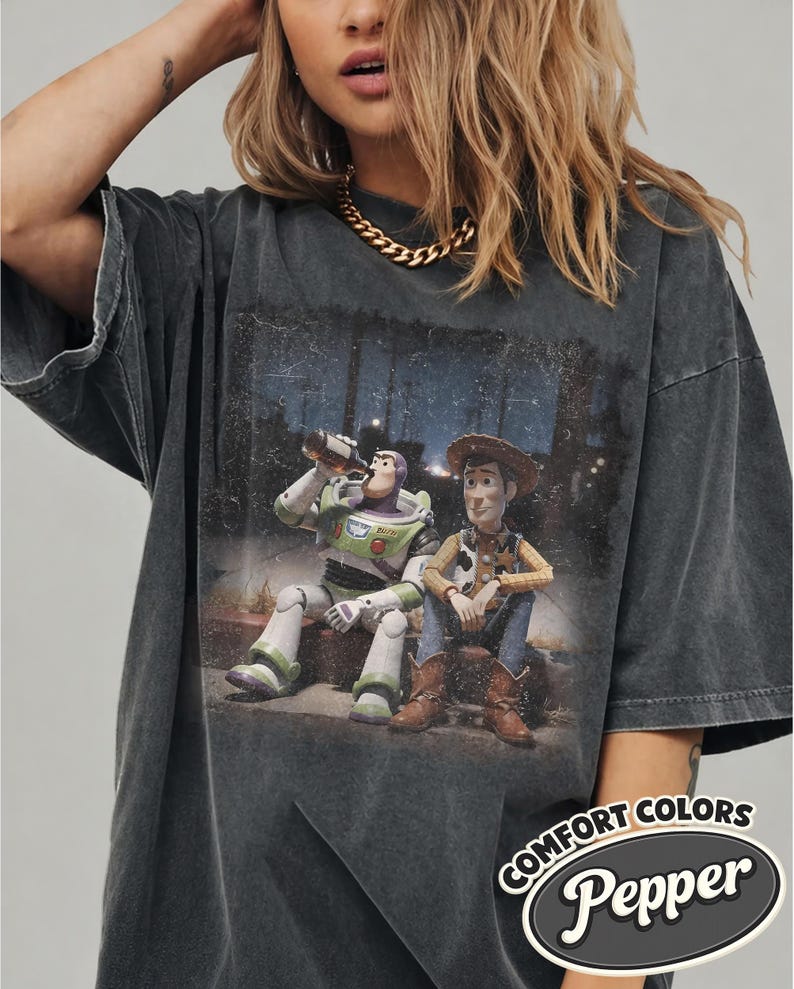 May include: Dark gray Comfort Colors t-shirt featuring a graphic of Buzz Lightyear and Woody from Toy Story. Buzz is drinking from a bottle, and Woody is sitting next to him. The shirt has a distressed look with the text "Comfort Colors Pepper".