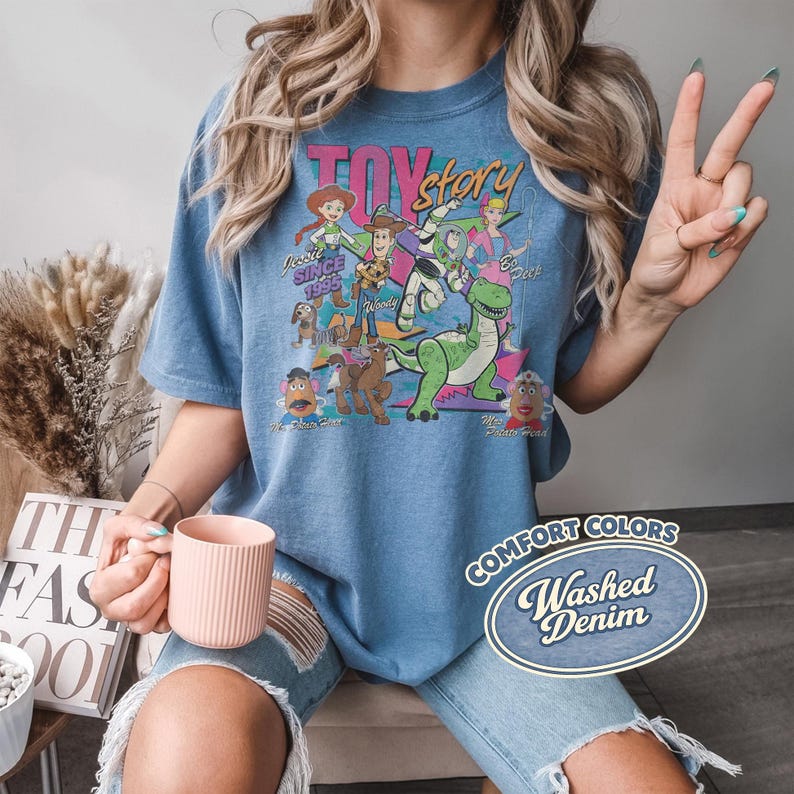 May include: Blue t-shirt featuring a graphic of Toy Story characters, including Woody, Buzz Lightyear, and Rex. The shirt has the text "TOY STORY" and "Since 1995". The shirt is labeled "Comfort Colors Washed Denim".