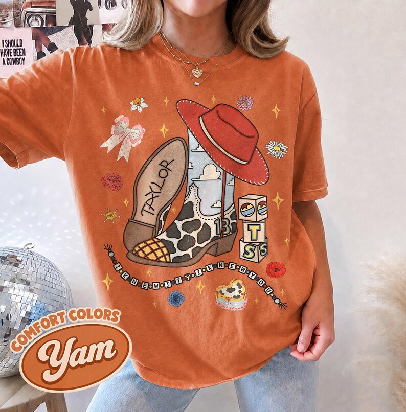 Peut inclure: Un t-shirt orange avec une impression graphique repr&eacute;sentant une botte de cowboy &agrave; motif de vache, un chapeau de cowboy rouge, un n&oelig;ud et trois blocs empil&eacute;s avec les lettres T, S et une ic&ocirc;ne de soleil. Le texte "TAYLOR" et "I KNEW IT" est pr&eacute;sent.