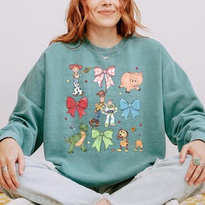 May include: A sage green crewneck sweatshirt featuring a graphic print of various animated characters including a cowgirl, a cowboy, a space ranger, a green dinosaur, a pig, and a dog, interspersed with large pink, red, blue, and green bows.