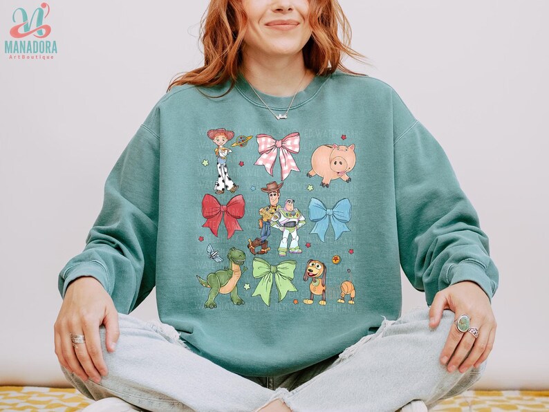 May include: A sage green crewneck sweatshirt featuring a graphic print of various animated characters including a cowgirl, a cowboy, a space ranger, a green dinosaur, a pig, and a dog, interspersed with large pink, red, blue, and green bows.