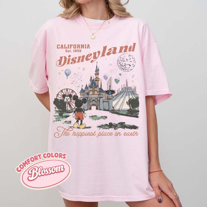 May include: Pink t-shirt with a Disneyland graphic. The design includes Mickey Mouse, a castle, and the text "California Disneyland Est. 1955." The shirt also features the phrase "The happiest place on earth."