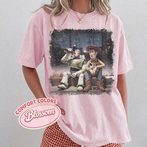 May include: Pink Comfort Colors t-shirt featuring a graphic of Buzz Lightyear and Woody from Toy Story. The characters are depicted sitting together. The shirt has the words "Comfort Colors Blossom" in a pink oval.