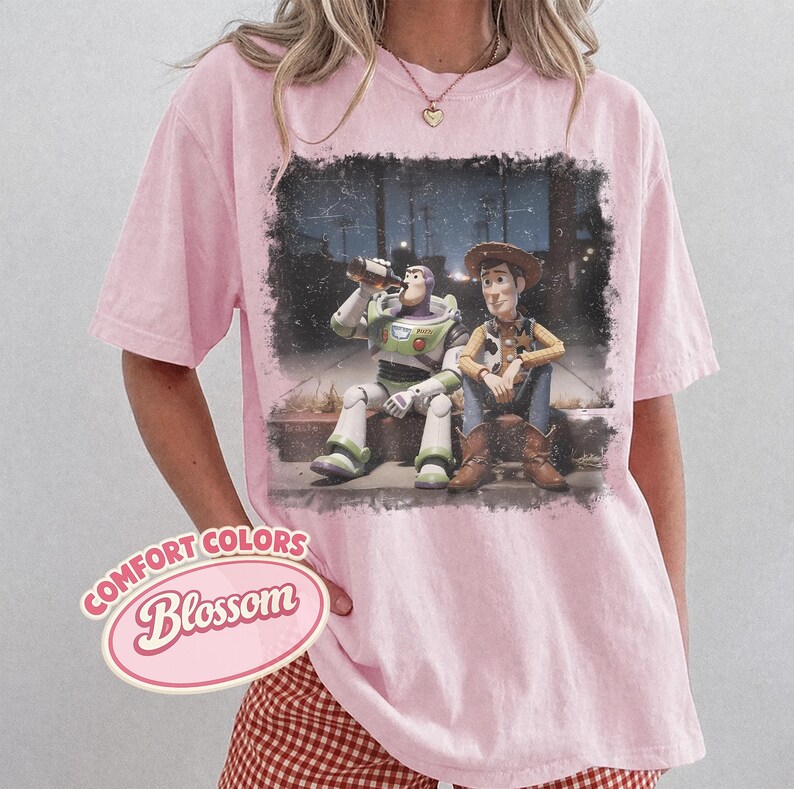 May include: Pink Comfort Colors t-shirt featuring a graphic of Buzz Lightyear and Woody from Toy Story. The characters are depicted sitting together. The shirt has the words "Comfort Colors Blossom" in a pink oval.