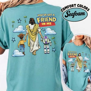 May include: A seafoam green t-shirt with a graphic of Jesus holding hands with cartoon characters. The text "YOU'VE GOT A FRIEND IN ME" is above the figures. The shirt also has the words "COMFORT COLORS" and "Seafoam" printed on it.