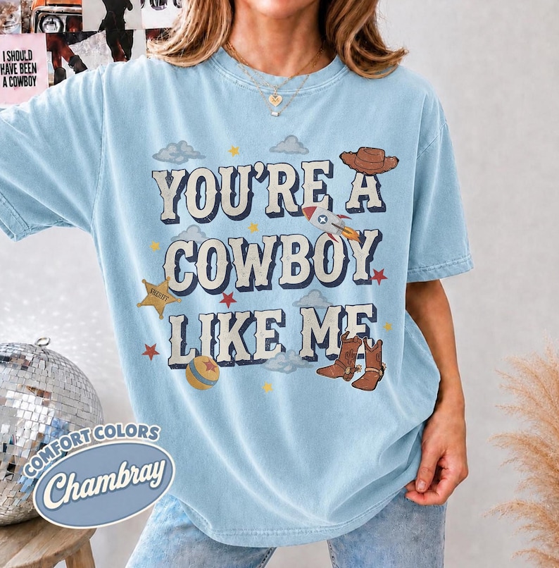 May include: A light blue short-sleeve t-shirt featuring the text "YOU'RE A COWBOY LIKE ME" in a dark blue font. The design includes illustrations of a brown cowboy hat, a rocket ship, a gold sheriff star, a ball, and brown cowboy boots.