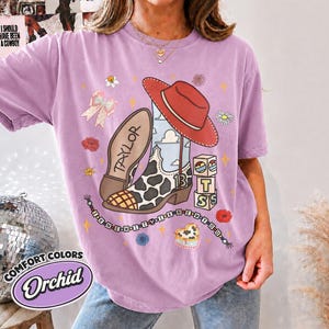Peut inclure: Un t-shirt couleur orchid&eacute;e avec un motif repr&eacute;sentant une botte de cow-boy &agrave; taches de vache, un chapeau rouge et des cubes formant le mot BTS. Le design inclut le texte TAYLOR, le nombre 13 et un collier de perles avec I KNEW IT I KNEW YOU.