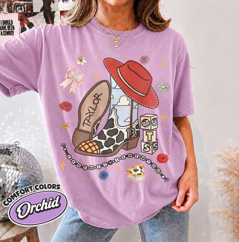 Peut inclure: Un t-shirt couleur orchid&eacute;e avec un motif repr&eacute;sentant une botte de cow-boy &agrave; taches de vache, un chapeau rouge et des cubes formant le mot BTS. Le design inclut le texte TAYLOR, le nombre 13 et un collier de perles avec I KNEW IT I KNEW YOU.
