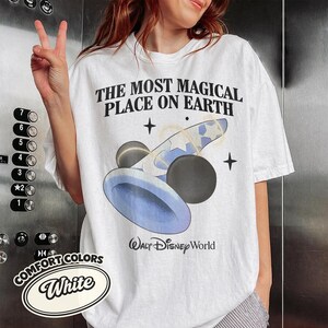 K&ouml;nnte beinhalten: Wei&szlig;es T-Shirt mit dem Text "THE MOST MAGICAL PLACE ON EARTH" und einer Grafik in Blau und Schwarz. Das Shirt tr&auml;gt auch die Worte "WALT Disney World" unter der Grafik. Das Shirt ist von Comfort Colors.