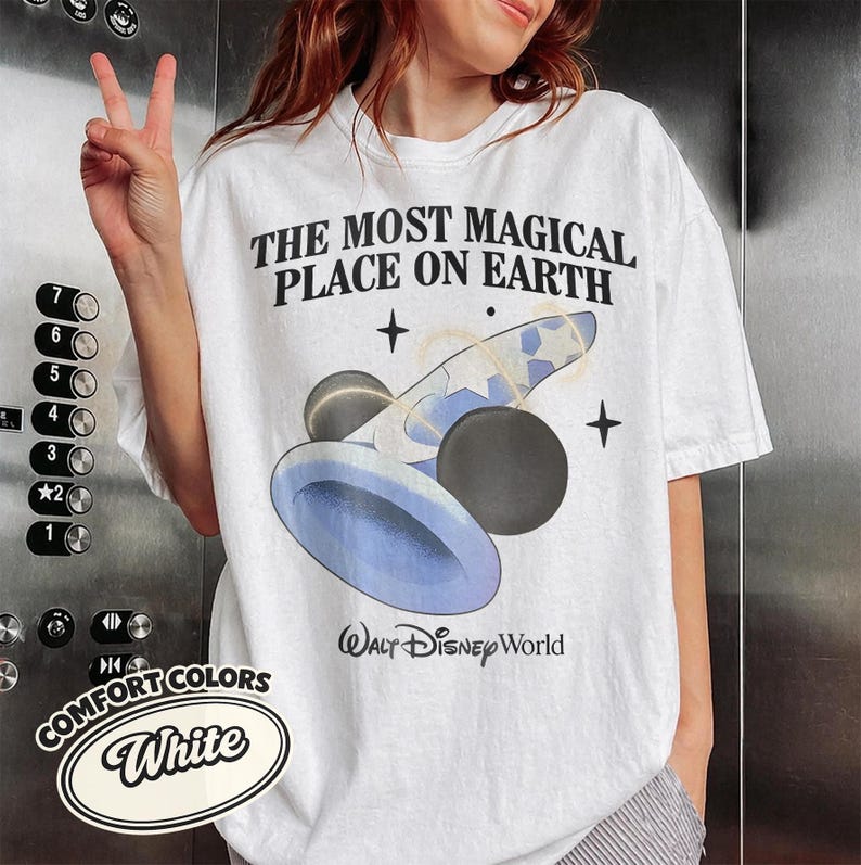 K&ouml;nnte beinhalten: Wei&szlig;es T-Shirt mit dem Text "THE MOST MAGICAL PLACE ON EARTH" und einer Grafik in Blau und Schwarz. Das Shirt tr&auml;gt auch die Worte "WALT Disney World" unter der Grafik. Das Shirt ist von Comfort Colors.