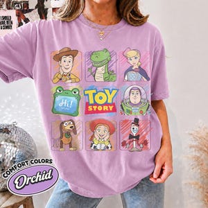 May include: A light purple t-shirt featuring a grid of Toy Story characters including Woody, Rex, Bo Peep, Buzz Lightyear, Slinky Dog, Jessie, and Forky, along with a green frog icon with the text Hi! and the Toy Story logo in the center.