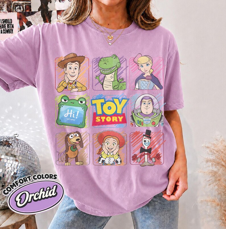 May include: A light purple t-shirt featuring a grid of Toy Story characters including Woody, Rex, Bo Peep, Buzz Lightyear, Slinky Dog, Jessie, and Forky, along with a green frog icon with the text Hi! and the Toy Story logo in the center.