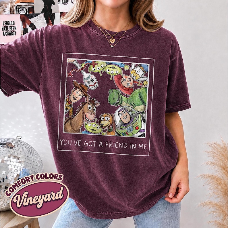 Puede incluir: Una camiseta Comfort Colors Vineyard de color granate con una ilustraci&oacute;n enmarcada de personajes de Toy Story. El texto "YOU'VE GOT A FRIEND IN ME" est&aacute; debajo de la imagen. La camiseta tambi&eacute;n tiene el texto "I SHOULD HAVE BEEN A COWBOY" en la parte superior izquierda.