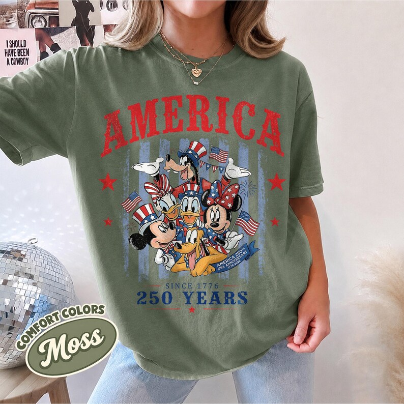 May include: An olive green short-sleeve t-shirt featuring a red graphic that reads AMERICA at the top. Below, cartoon characters wear patriotic hats and accessories with American flags. Text at the bottom reads SINCE 1776 250 YEARS.
