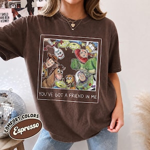 Puede incluir: Camiseta color espresso con un gr&aacute;fico enmarcado de personajes animados, incluyendo un vaquero, un guardabosques espacial y extraterrestres verdes. El texto "YOU'VE GOT A FRIEND IN ME" est&aacute; impreso debajo. La camiseta es parte de la colecci&oacute;n Comfort Colors.