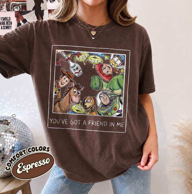 Puede incluir: Camiseta color espresso con un gr&aacute;fico enmarcado de personajes animados, incluyendo un vaquero, un guardabosques espacial y extraterrestres verdes. El texto "YOU'VE GOT A FRIEND IN ME" est&aacute; impreso debajo. La camiseta es parte de la colecci&oacute;n Comfort Colors.