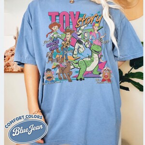 May include: Blue short-sleeved t-shirt with a graphic featuring characters from the movie Toy Story. The design includes Jessie, Woody, Buzz Lightyear, and others, with the text "TOY STORY" and "SINCE 1995". The shirt is labeled "COMFORT COLORS Blue Jean".