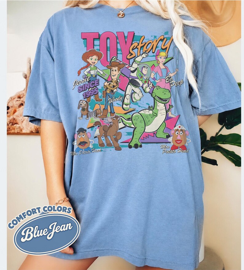 May include: Blue short-sleeved t-shirt with a graphic featuring characters from the movie Toy Story. The design includes Jessie, Woody, Buzz Lightyear, and others, with the text "TOY STORY" and "SINCE 1995". The shirt is labeled "COMFORT COLORS Blue Jean".