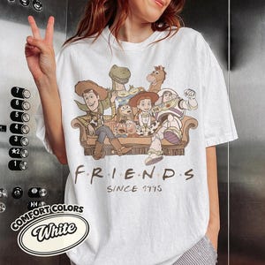 Puede incluir: Camiseta blanca con un gr&aacute;fico de personajes de Toy Story sentados en un sof&aacute; marr&oacute;n. La palabra "FRIENDS" est&aacute; impresa en negro, con "SINCE 1995" debajo. La camiseta tambi&eacute;n tiene un logotipo de "COMFORT COLORS".