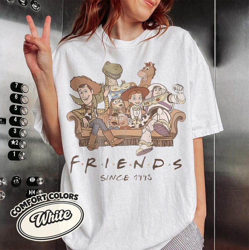 Puede incluir: Camiseta blanca con un gr&aacute;fico de personajes de Toy Story sentados en un sof&aacute; marr&oacute;n. La palabra "FRIENDS" est&aacute; impresa en negro, con "SINCE 1995" debajo. La camiseta tambi&eacute;n tiene un logotipo de "COMFORT COLORS".