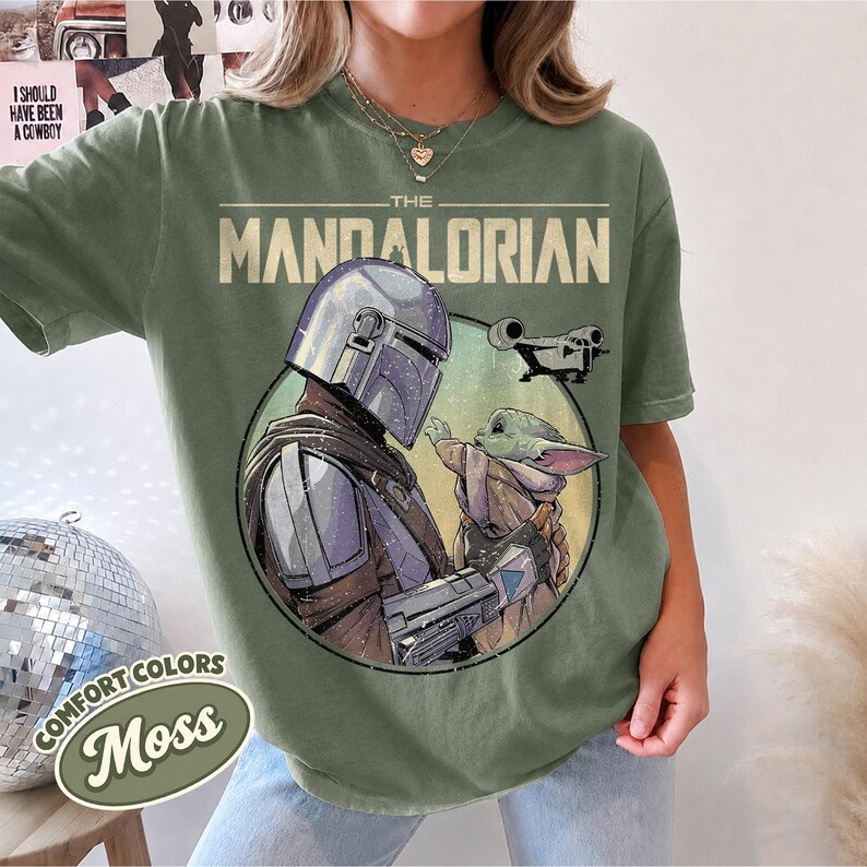 Puede incluir: Una camiseta de manga corta color verde musgo con un estampado gr&aacute;fico de The Mandalorian y Grogu dentro de un marco circular. El texto THE MANDALORIAN est&aacute; impreso en una fuente clara y llamativa sobre los personajes, con una peque&ntilde;a nave.