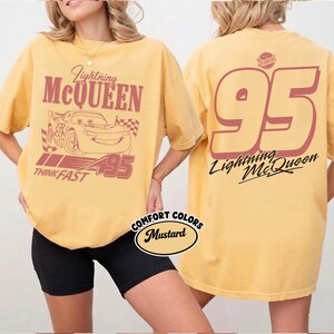 May include: Mustard-colored t-shirt featuring Lightning McQueen from the movie Cars. The front graphic includes the character, the number 95, and the words "Lightning McQueen" and "Think Fast." The back of the shirt displays a large number 95 and the character's name.