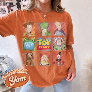 May include: An orange short-sleeve t-shirt featuring a grid of Toy Story characters including Woody, Rex, Bo Peep, a green frog with the text Hi!, Buzz Lightyear, Slinky Dog, Jessie, and Forky, with the Toy Story logo in the center.
