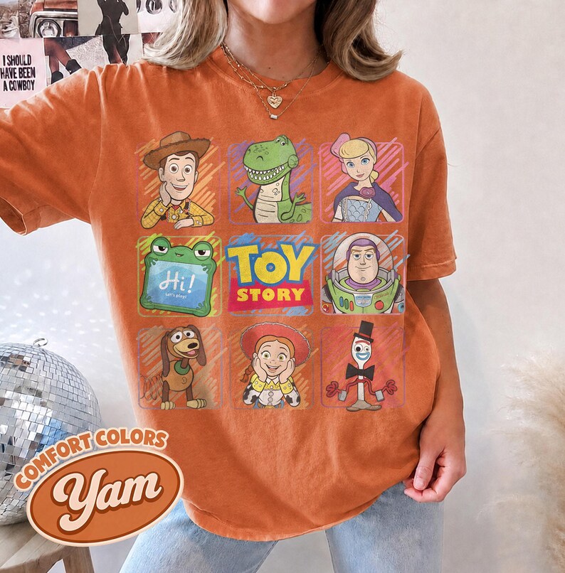 May include: An orange short-sleeve t-shirt featuring a grid of Toy Story characters including Woody, Rex, Bo Peep, a green frog with the text Hi!, Buzz Lightyear, Slinky Dog, Jessie, and Forky, with the Toy Story logo in the center.