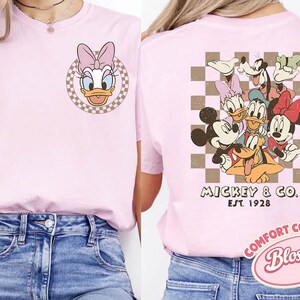 May include: A light pink t-shirt featuring Daisy Duck on the front and a group of Disney characters on the back. The front has a circular design with Daisy Duck's face. The back has Mickey & Co. Est. 1928.