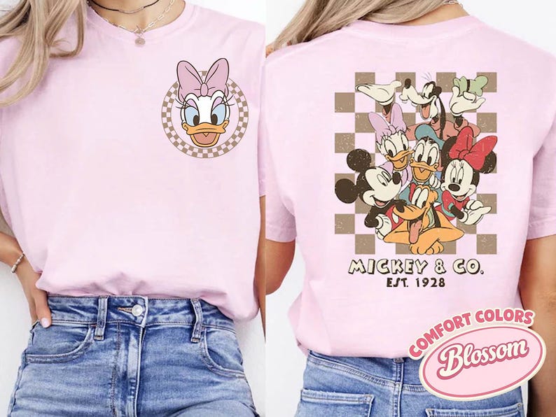 May include: A light pink t-shirt featuring Daisy Duck on the front and a group of Disney characters on the back. The front has a circular design with Daisy Duck's face. The back has Mickey & Co. Est. 1928.