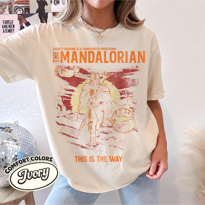 May include: An ivory short-sleeve t-shirt featuring a distressed graphic print of The Mandalorian. The text reads, "Bounty hunting is a complicated profession... The Mandalorian" and "This is the way" in orange, with a yellow sun background.