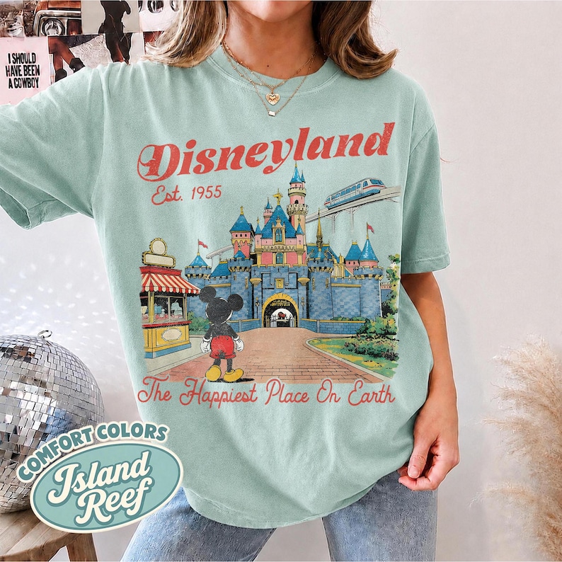 May include: A light green short-sleeve t-shirt featuring a graphic of a castle, a monorail, and Mickey Mouse. The text reads Disneyland Est. 1955 and The Happiest Place On Earth in red script. The design includes a red and white striped kiosk.
