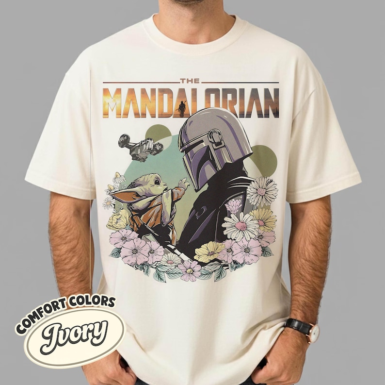 May include: Ivory-colored t-shirt featuring a graphic of The Mandalorian and Grogu surrounded by flowers. The design includes the text "THE MANDALORIAN" in a sunset-like font, with a spaceship in the background. The shirt is labeled "COMFORT COLORS Ivory".