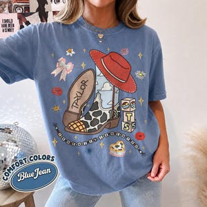 Peut inclure: Un t-shirt bleu jean avec une impression graphique repr&eacute;sentant une botte de cow-boy &agrave; motif de vache, un chapeau rouge, le nombre 13, des blocs avec les lettres T, S et un collier de perles avec le texte I KNEW IT I KNEW YOU.