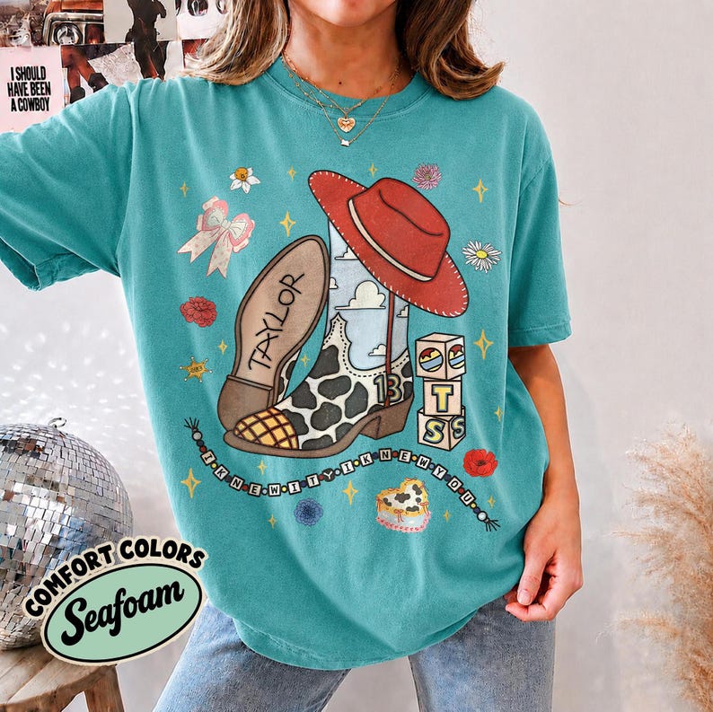 Peut inclure: Un t-shirt &agrave; manches courtes bleu sarcelle avec une impression graphique d'une botte de cow-boy &agrave; motifs de vache, un chapeau de cow-boy rouge, des blocs avec le texte T S, et le texte TAYLOR et I KNEW IT I KNEW YOU, avec des fleurs et &eacute;toiles.
