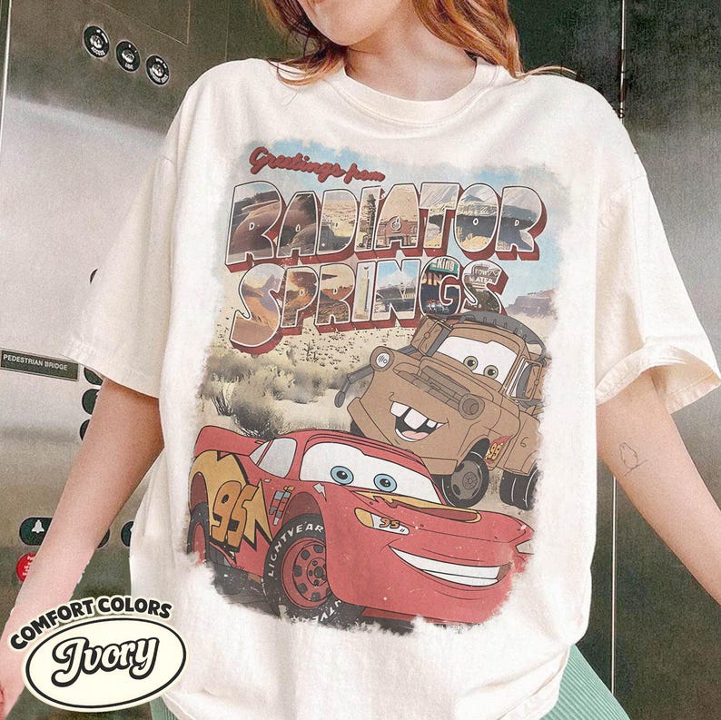 May include: Ivory-colored t-shirt featuring a graphic of Lightning McQueen and Mater from the movie Cars, with the text "Greetings from Radiator Springs." The shirt is from Comfort Colors.