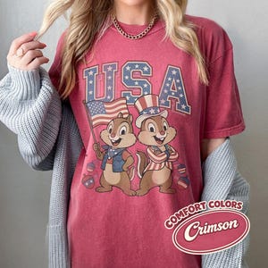 May include: Crimson-colored t-shirt featuring the word "USA" in a blue and white star-filled font. Two cartoon chipmunks are depicted, one holding a US flag and the other wearing a patriotic hat. The shirt has a vintage look.