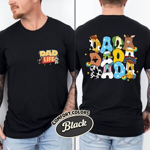 May include: Black t-shirt with "DAD LIFE" and Toy Story characters on the front. The back features the word "DAD" in colorful, patterned letters with Toy Story character designs. The shirt is labeled "Comfort Colors Black".