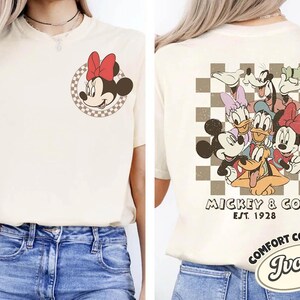 May include: Ivory-colored t-shirt featuring Minnie Mouse with a red bow on the front and a group of Disney characters on the back. The back design includes Mickey Mouse, Donald Duck, Goofy, and Daisy Duck. The shirt has the text "Mickey & Co. Est. 1928".