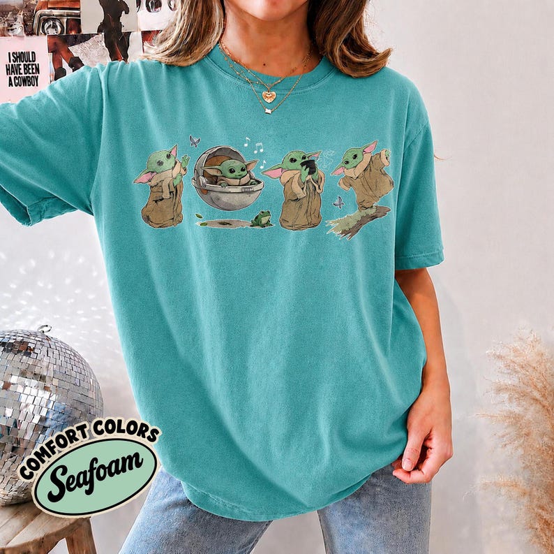 May include: A teal short-sleeve t-shirt featuring a graphic print of four small green creatures in brown robes. The design shows the characters in various poses, including sitting in a floating pod, holding a small frog, and standing.
