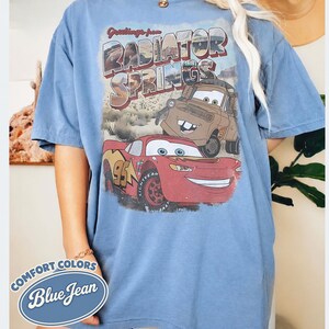 May include: Blue short-sleeve t-shirt with a graphic of the Cars characters Lightning McQueen and Mater, and the text "Greetings from Radiator Springs." The shirt also has the text "Comfort Colors Blue Jean."