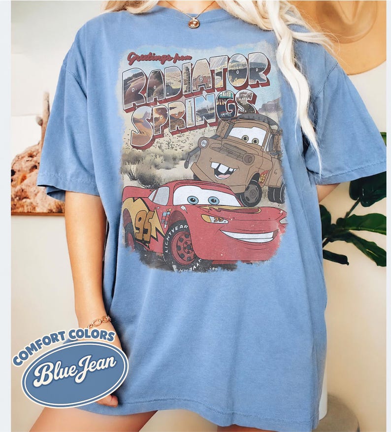 May include: Blue short-sleeve t-shirt with a graphic of the Cars characters Lightning McQueen and Mater, and the text "Greetings from Radiator Springs." The shirt also has the text "Comfort Colors Blue Jean."